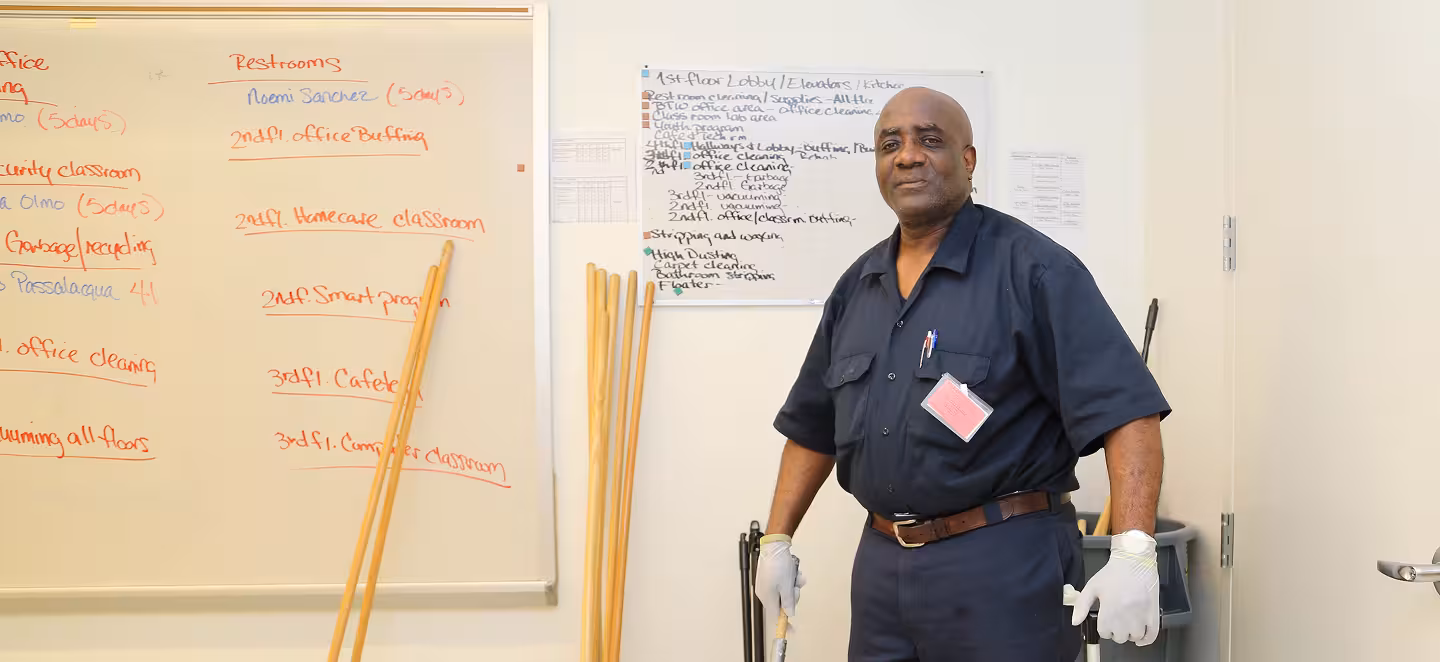 Custodial workers in overalls and yellow gloves vacuuming carpet and cleaning floor-to-ceiling windows in a bright office.