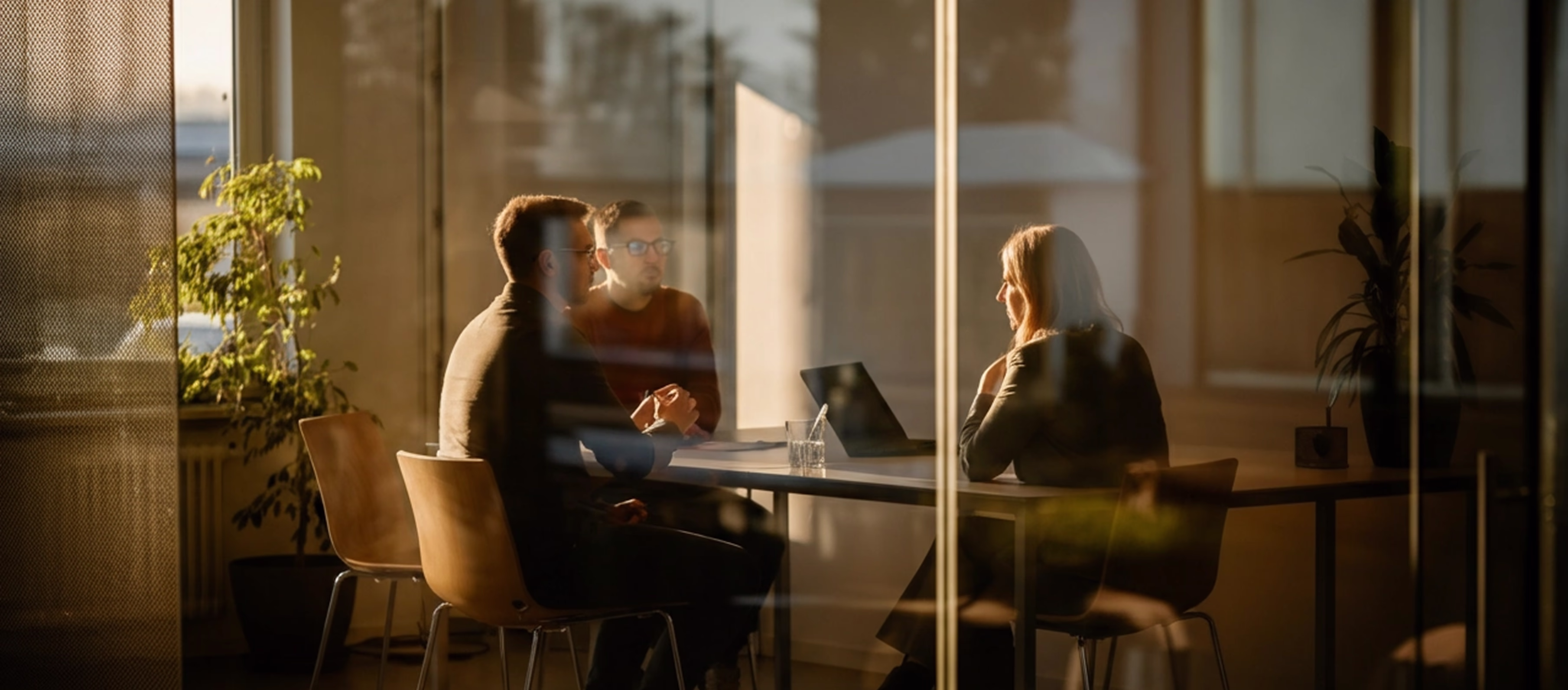Three people seated around a table in a sunlit office, engaged in a discussion with a laptop and glass of water on the table.