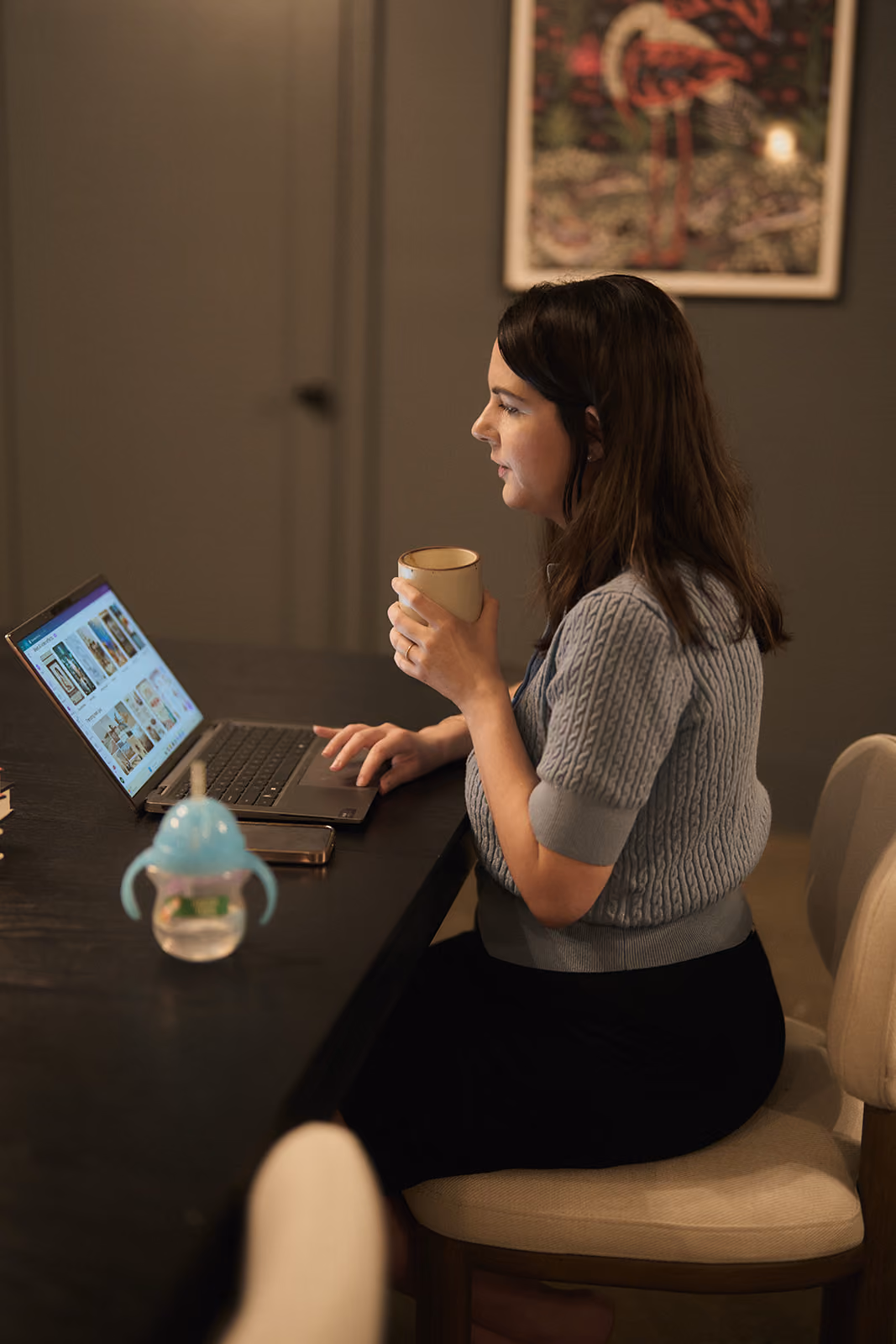 Woman in gray sweater sitting at table using laptop and holding a beige mug.