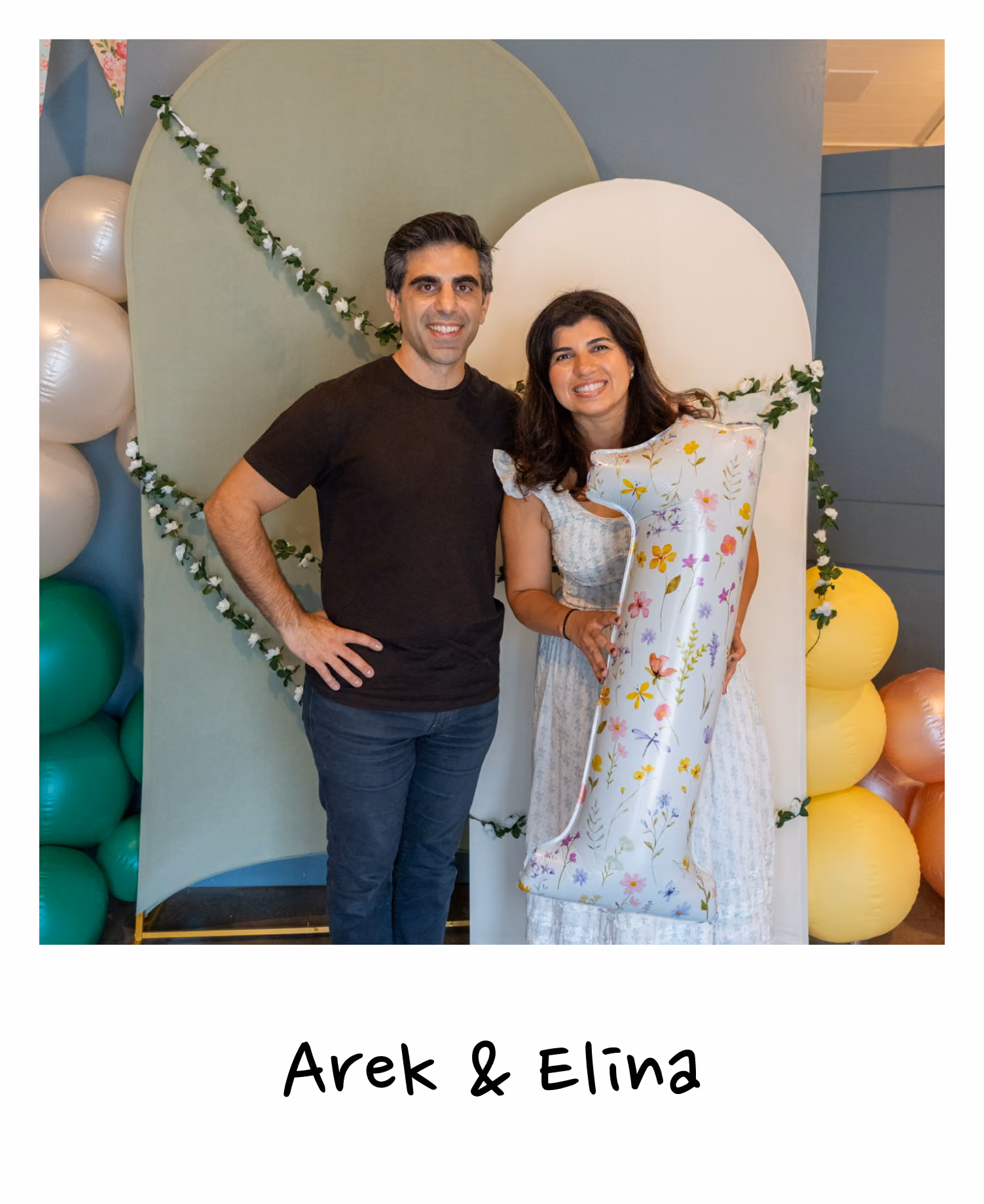 A man with dark hair wearing a black t-shirt and dark jeans stands next to a woman with long dark hair wearing a white dress, who is holding a large silver balloon shaped like the number one with colorful floral prints, against a decorative backdrop with green and white panels and balloons.