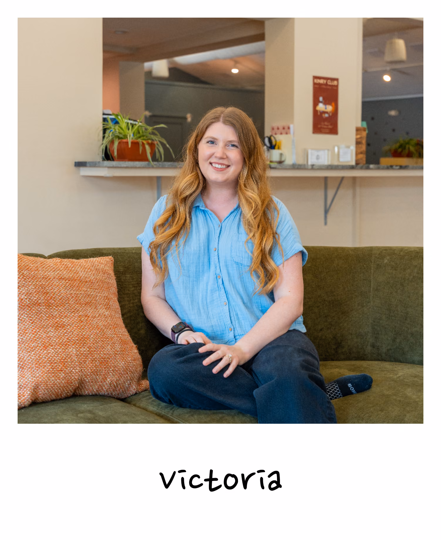A smiling woman with long wavy light brown hair wearing a light blue short-sleeve shirt and dark pants sitting cross-legged on a green couch with an orange pillow, in a cozy indoor space with plants and a kitchen counter in the background.