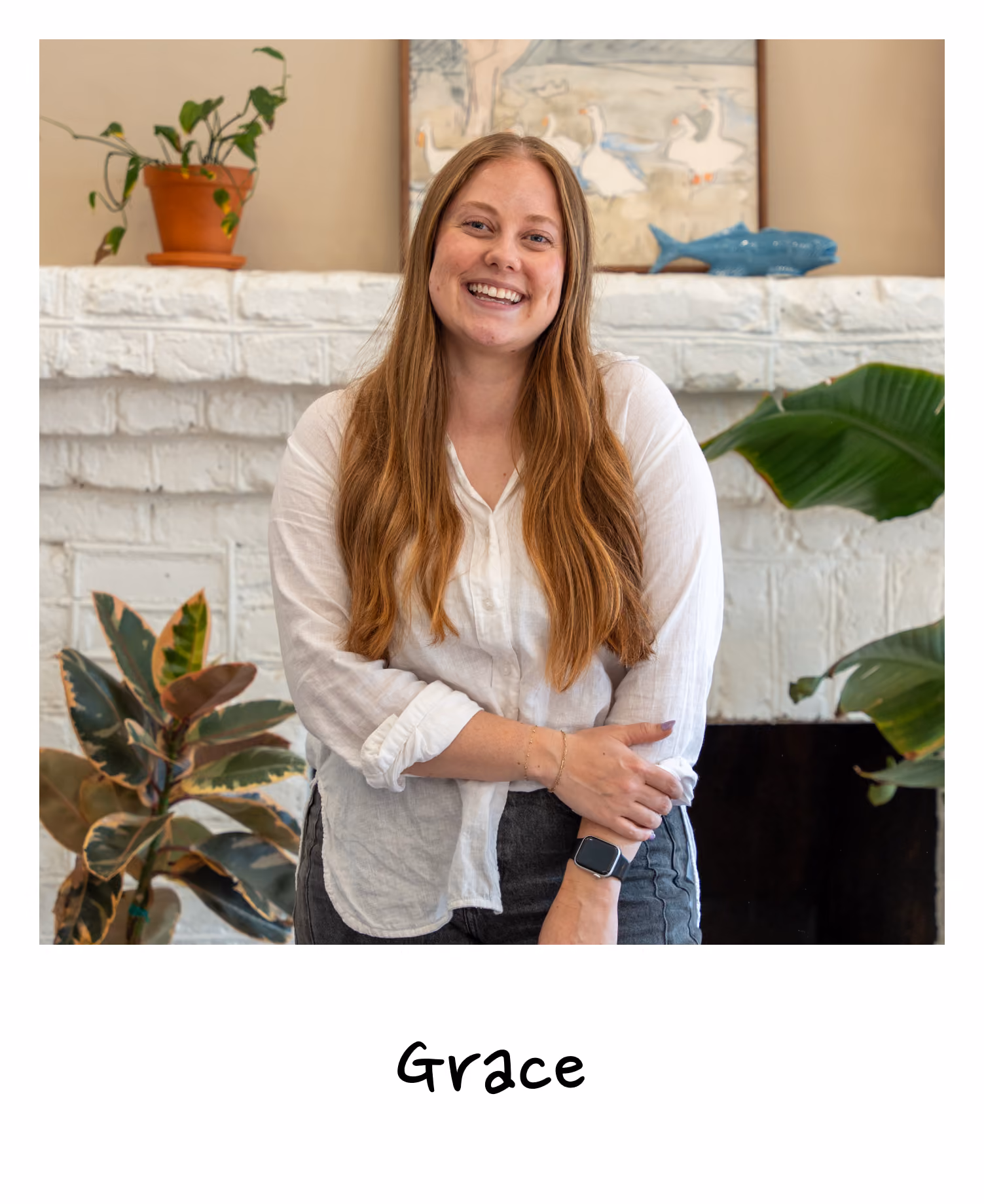 Smiling young woman with long light brown hair wearing a white shirt and dark pants, standing indoors in front of a white brick fireplace decorated with plants and artwork.