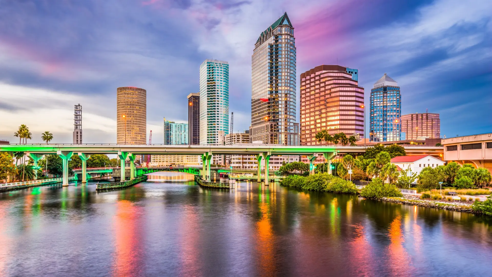 City skyline at dusk with high-rise buildings, a bridge over river, and colorful reflections in water.