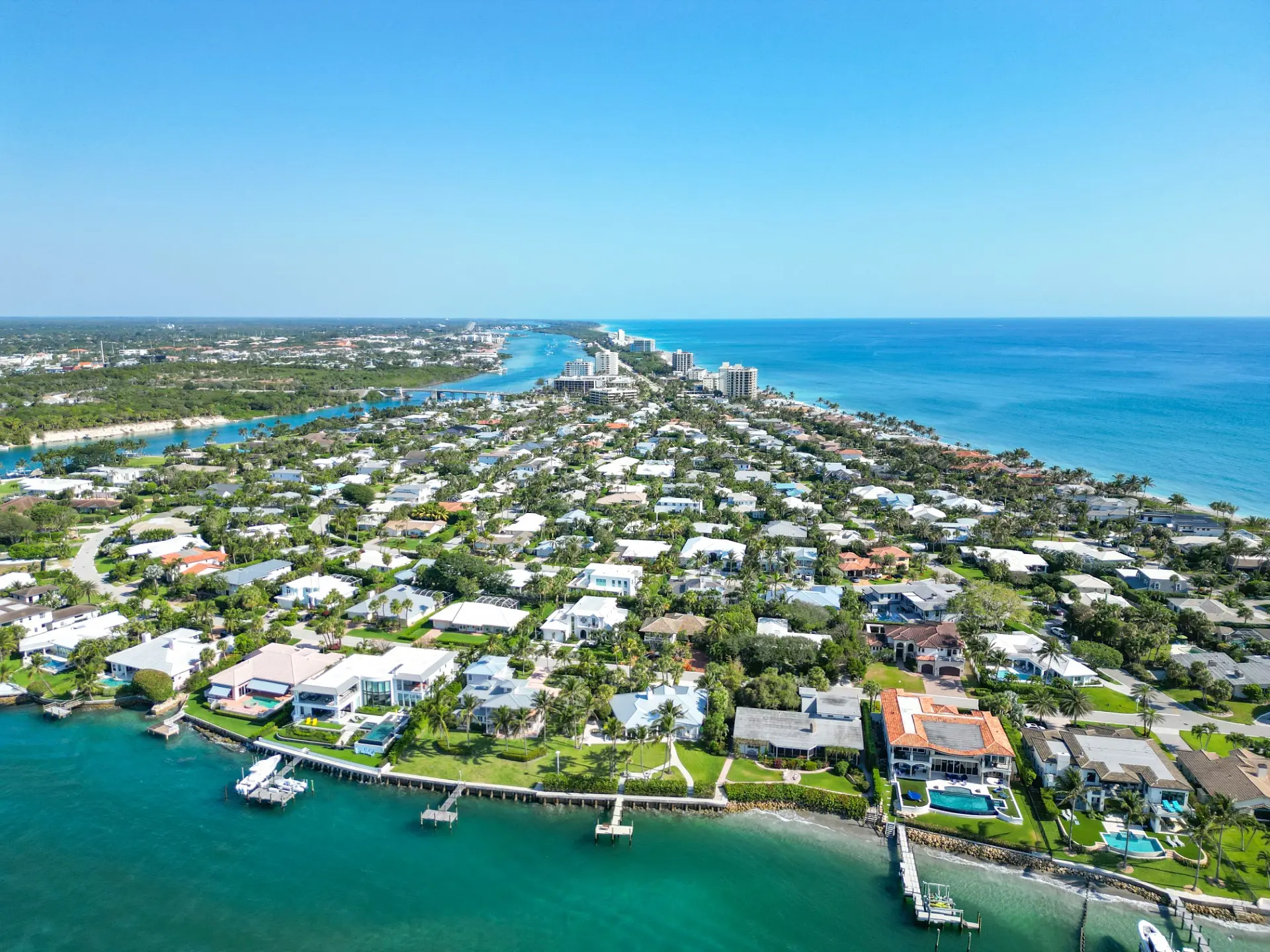 Aerial view of coastal residential area with houses, docks, greenery, and ocean under a clear blue sky.