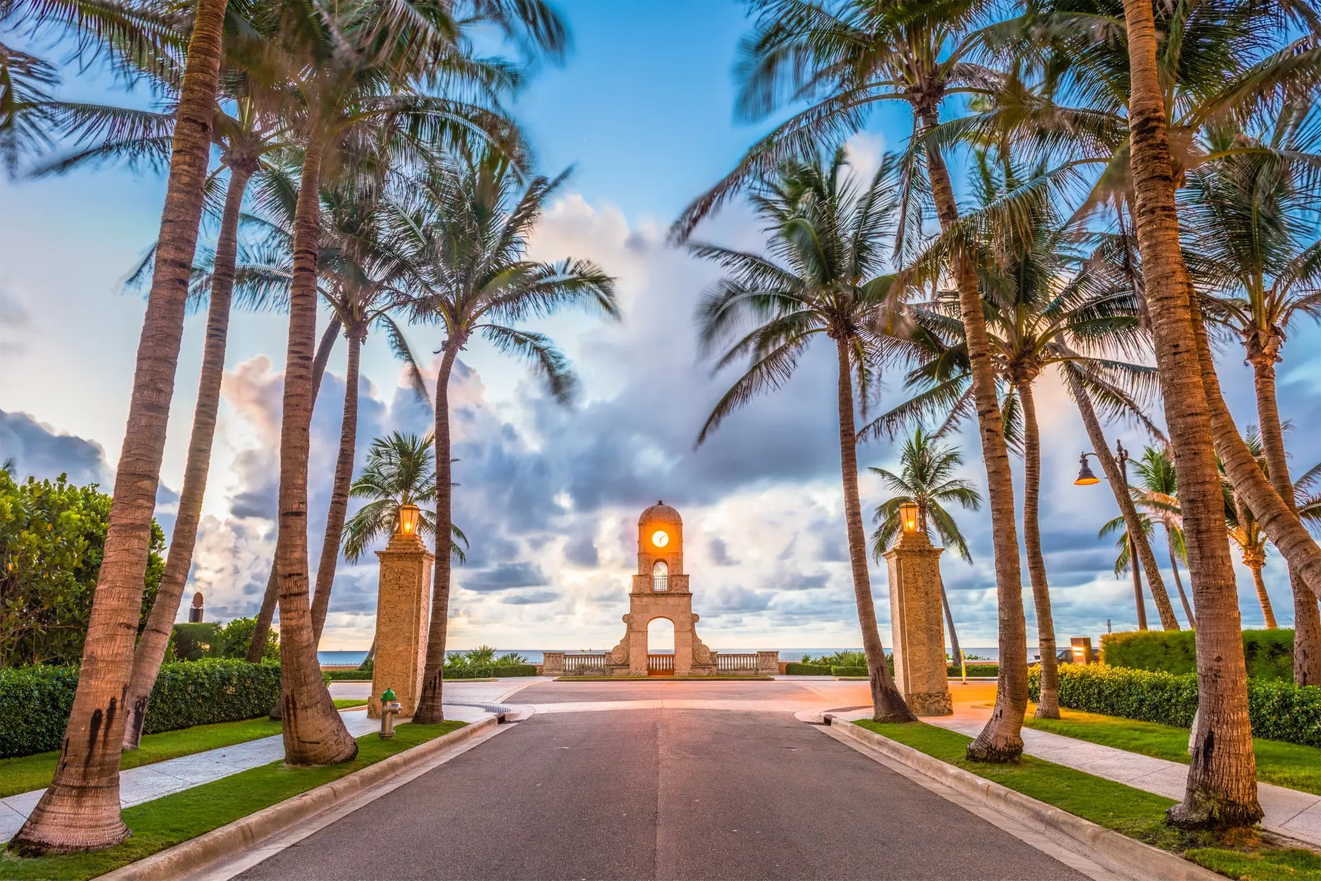 Palm-lined road leading to stone clock tower with glowing lamps at sunset by ocean.