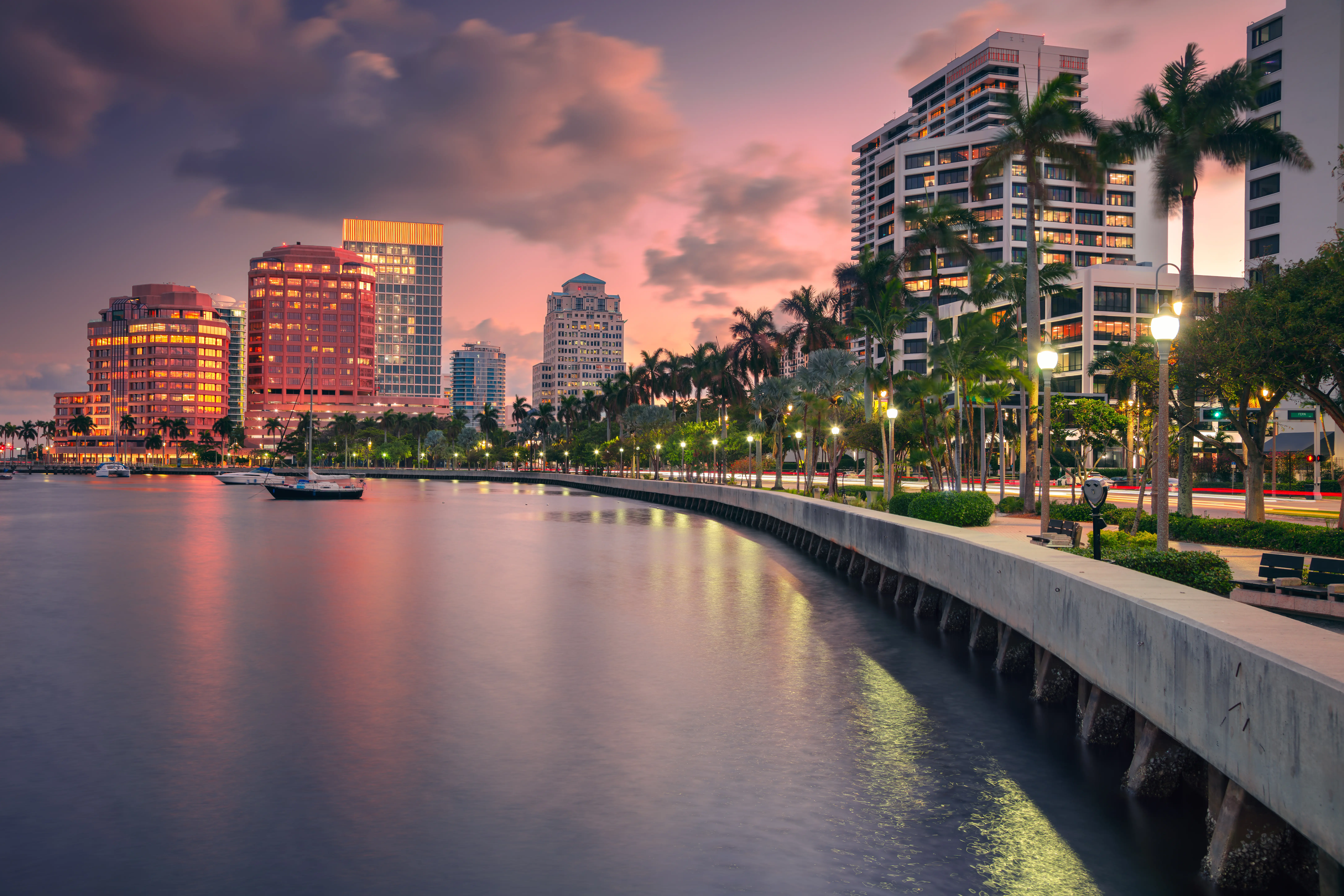 Waterfront cityscape at sunset with lit buildings, palm trees, and boats in calm water.