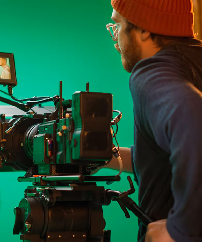 Man wearing glasses and an orange beanie operating a professional video camera on a tripod with a green screen background.