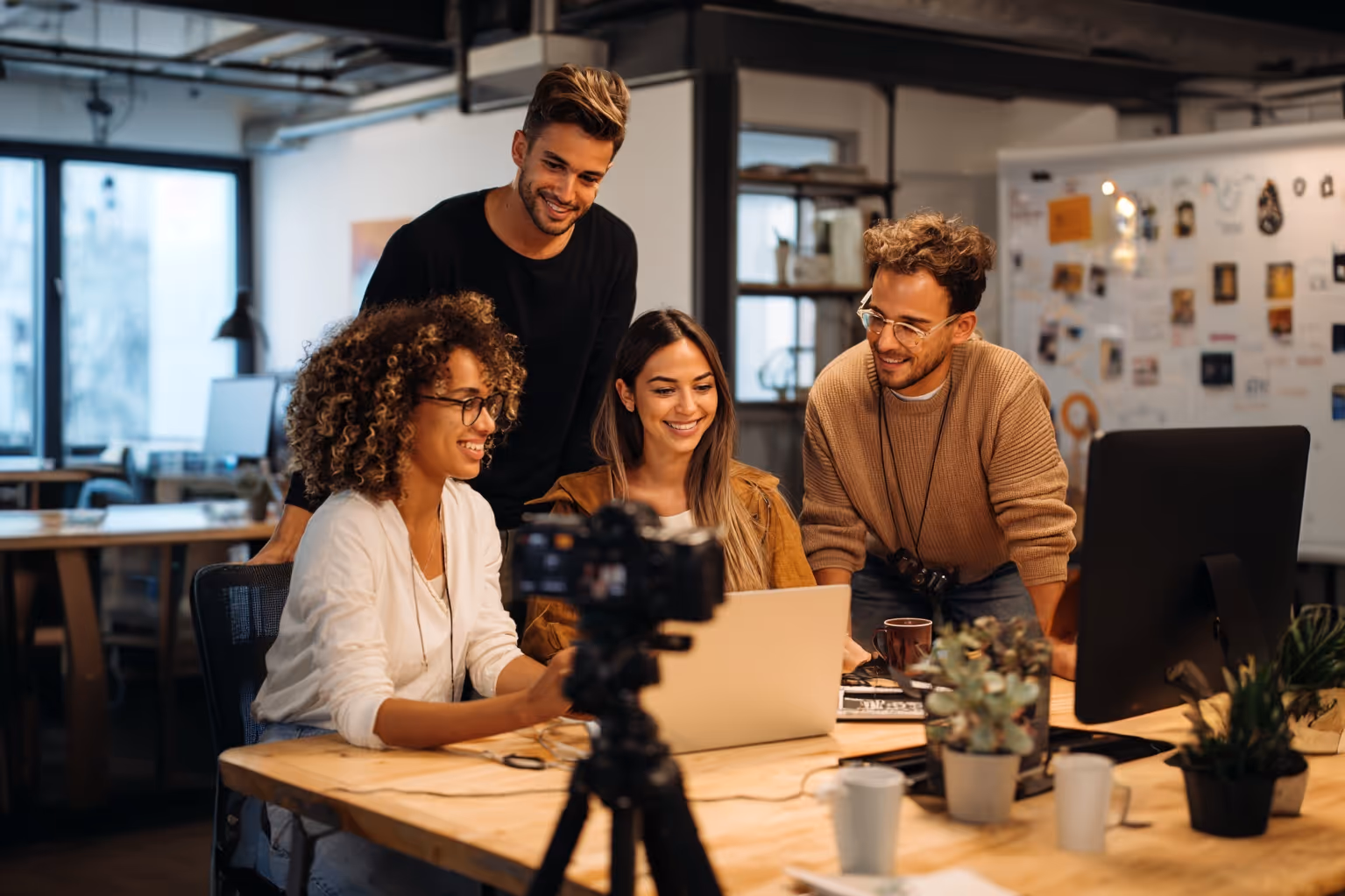 Four young professionals collaborating and smiling around a laptop in a modern office with a camera on a tripod in the foreground.