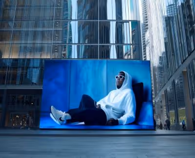 Large digital billboard in urban plaza showing a person in white hoodie and sunglasses reclining against blue background.