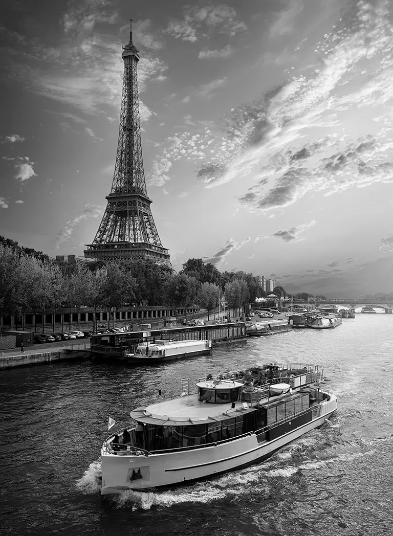 Image en noir et blanc de la Tour Eiffel au bord de la Seine à Paris. Un bateau mouche navigue au premier plan et le ciel est rempli de nuages épars. Ambiance Solamor.