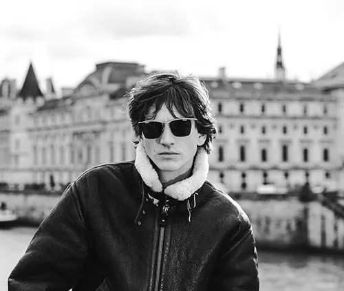 Un homme portant des lunettes de soleil Solamor et une veste en cuir avec un col en peau de mouton se tient à l'extérieur, des bâtiments historiques encadrant la scène. La photo en noir et blanc capture une allure intemporelle avec en arrière plan, la Conciergerie de Paris.