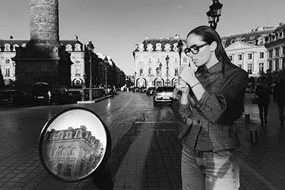 Une femme portant des lunettes allume une cigarette dans la rue, place Vendôme à Paris. Vêtue d'une veste et d'un jean. Un miroir circulaire de scooter reflète les bâtiments voisins, capturant l'essence de l'architecture historique. Ses lunettes élégantes ajoutant un air de sophistication à cette scène en noir et blanc.