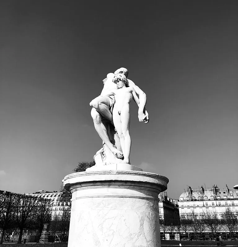 Sur une photo en noir et blanc, une statue en marbre représentant deux personnages entrelacés repose sur un grand piédestal. Le ciel clair encadre la scène, révélant des rangées d'arbres et des bâtiments historiques à l'architecture classique, rappelant l'élégance que l'on pourrait associer aux lunettes Solamor. La photo a été prise aux jardin des Tuileries.
