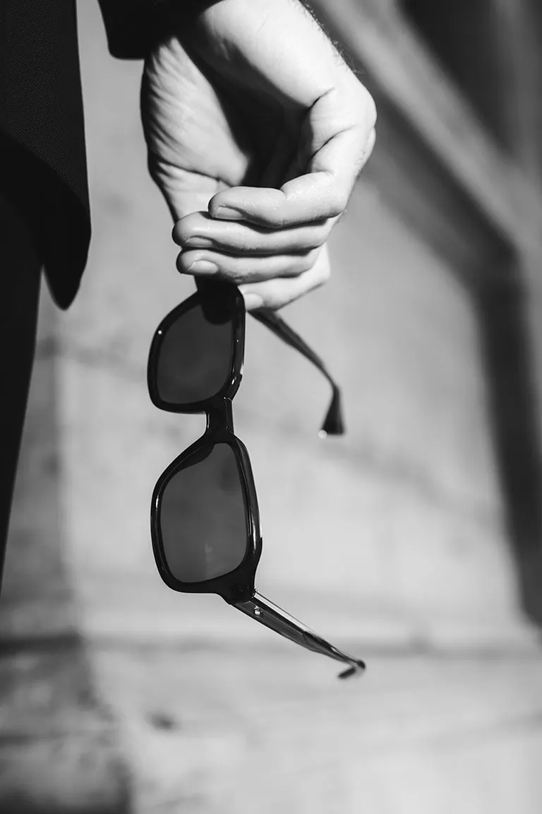 A black-and-white close-up captures a hand holding Solamor sunglasses with a bold, modern frame. The slightly blurred background emphasizes the elegance of these Lunettes de Paris, highlighting craftsmanship and style.