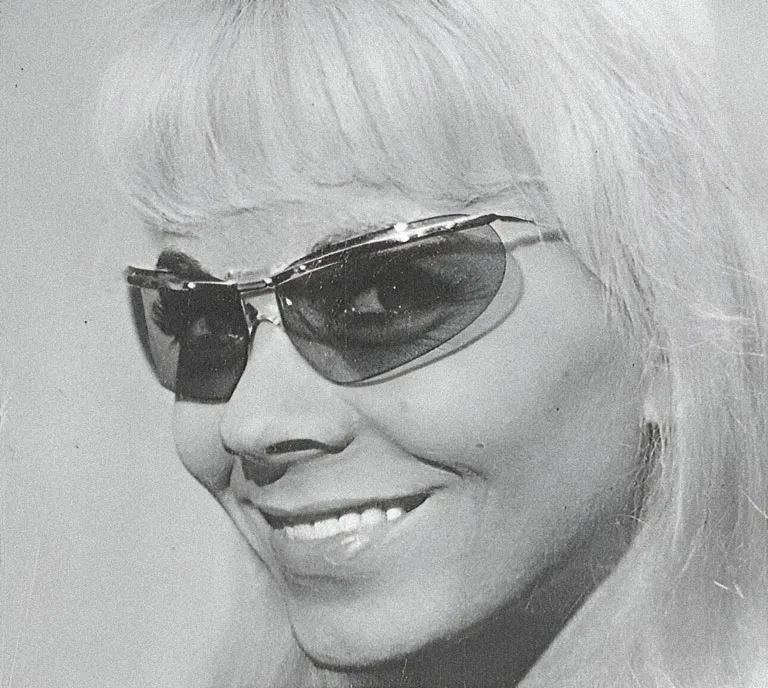 Une femme aux cheveux blonds, avec une frange, sourit tout en portant des lunettes de soleil fumées, le modèle Sport 1000 crée en 1961. La photo est en noir et blanc, ce qui met en valeur le style vintage des lunettes.
