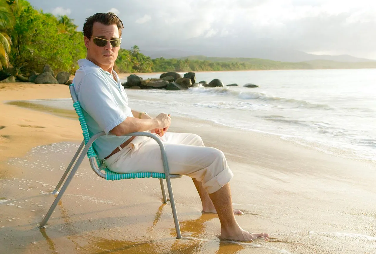 hnny Depp wearing Solamor sunglasses, Sport 1000 model, on the set of the film "The Rum Diary." He sits on an outdoor chair by the beach, feet in the water, dressed entirely in white.
