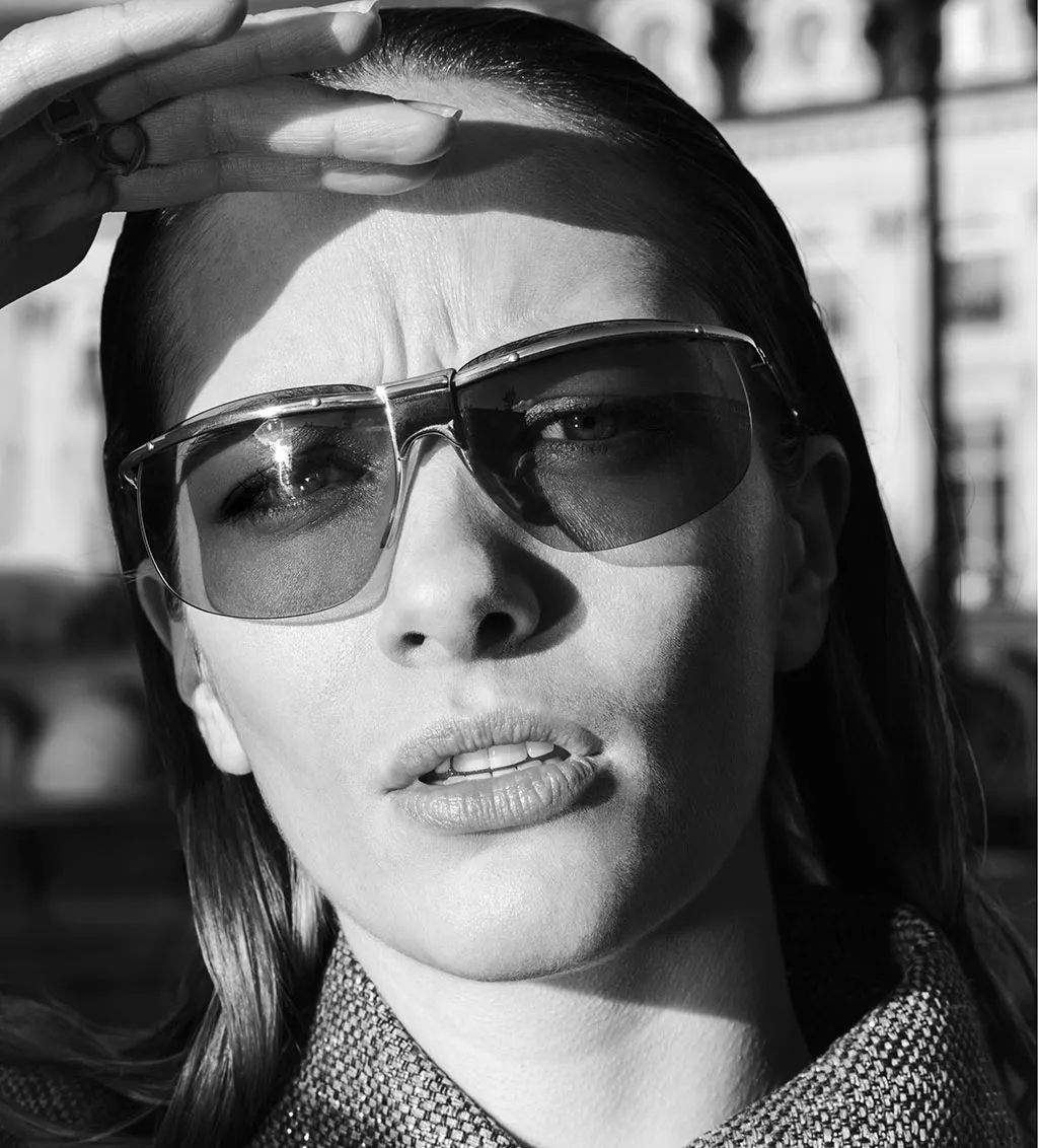 Black-and-white close-up of a woman wearing Solamor sunglasses, looking upward with one hand partially shielding their eyes from the sun. The blurred background hints at architecture, evoking the atmosphere of the french brand Solamor.