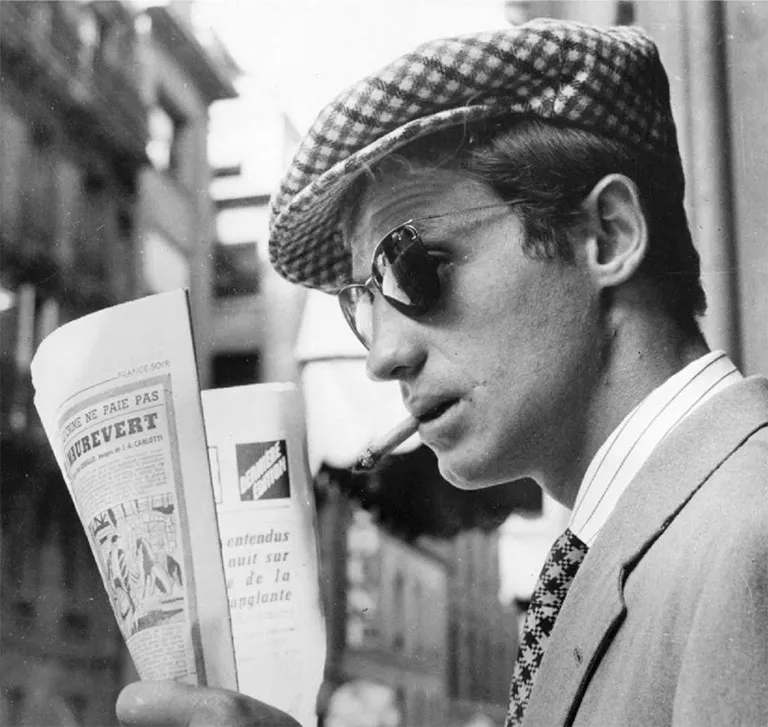 Jean-Paul Belmondo on the set of "Breathless" or " A bout de souffle" by Jean-Luc Godard. The actor is in profile, cigarette in mouth, holding a newspaper, dressed in a suit and tie, wearing Solamor glasses.