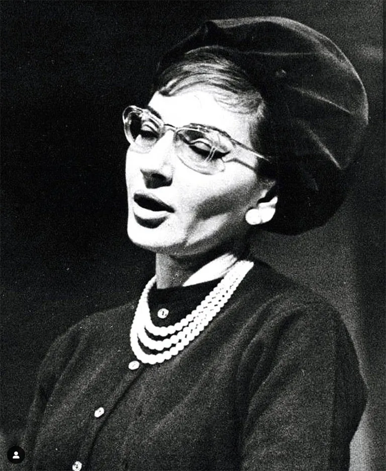 Maria Callas, the famous Greek opera singer, wears Solamor eyeglasses. She is dressed in a black cardigan and a three-strand pearl necklace. Her hair is styled under a black hat.