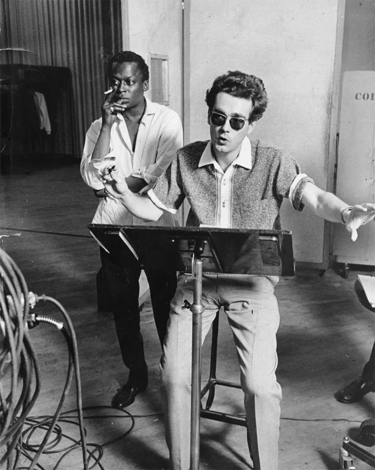 Michel Legrand — renowned musician, composer, jazz pianist, and singer — in rehearsal, making a hand gesture as if beating the music. A music stand stands before him, and a man smokes in the background. He wears a vintage pair of Solamor glasses.