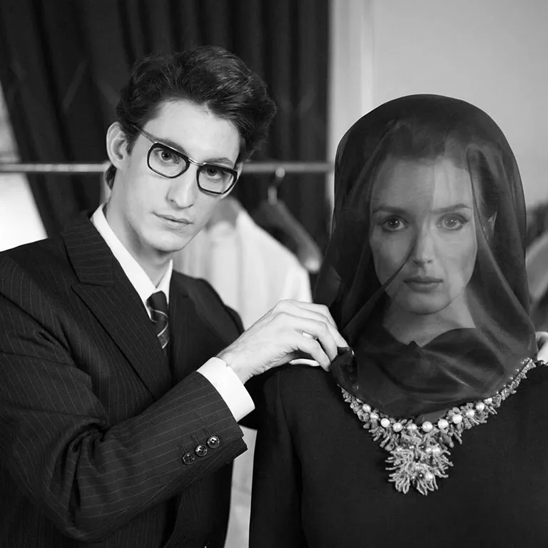 Pierre Niney and Charlotte Le Bon on the set of the film "Saint Laurent," a biopic about the life of fashion designer Yves Saint Laurent. Here, the actor wears vintage Solamor glasses while placing a necklace around Charlotte Le Bon’s neck.