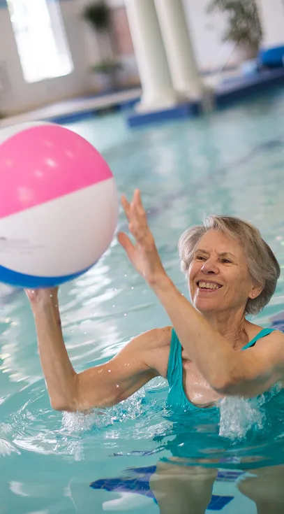 Smiling older woman playing with a beach ball in a bright indoor pool, enjoying a fun and active moment in the water.
