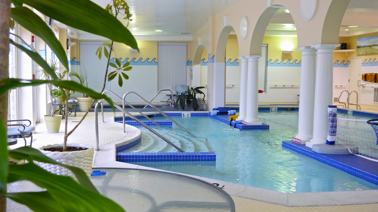 A bright indoor pool area featuring arched columns, tiled walls, and a curved pool with railings and built-in seating. Lush green plants and natural light create a relaxing, resort-style atmosphere.