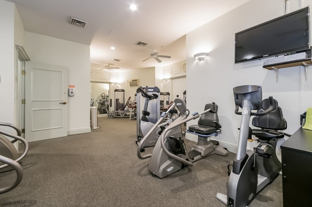 Well-lit fitness room featuring stationary bikes, elliptical machines, and strength training equipment. The space includes a wall-mounted TV, ceiling fans, and mirrors to support a comfortable and functional workout environment.
