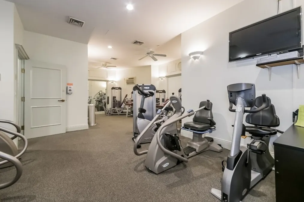 Well-lit fitness room featuring stationary bikes, elliptical machines, and strength training equipment. The space includes a wall-mounted TV, ceiling fans, and mirrors to support a comfortable and functional workout environment.
