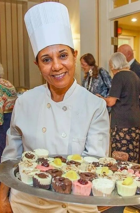 A pastry chef in a white uniform and tall chef hat holds a tray filled with colorful mini desserts. Residents in the background enjoy a social gathering, creating a warm and festive atmosphere.