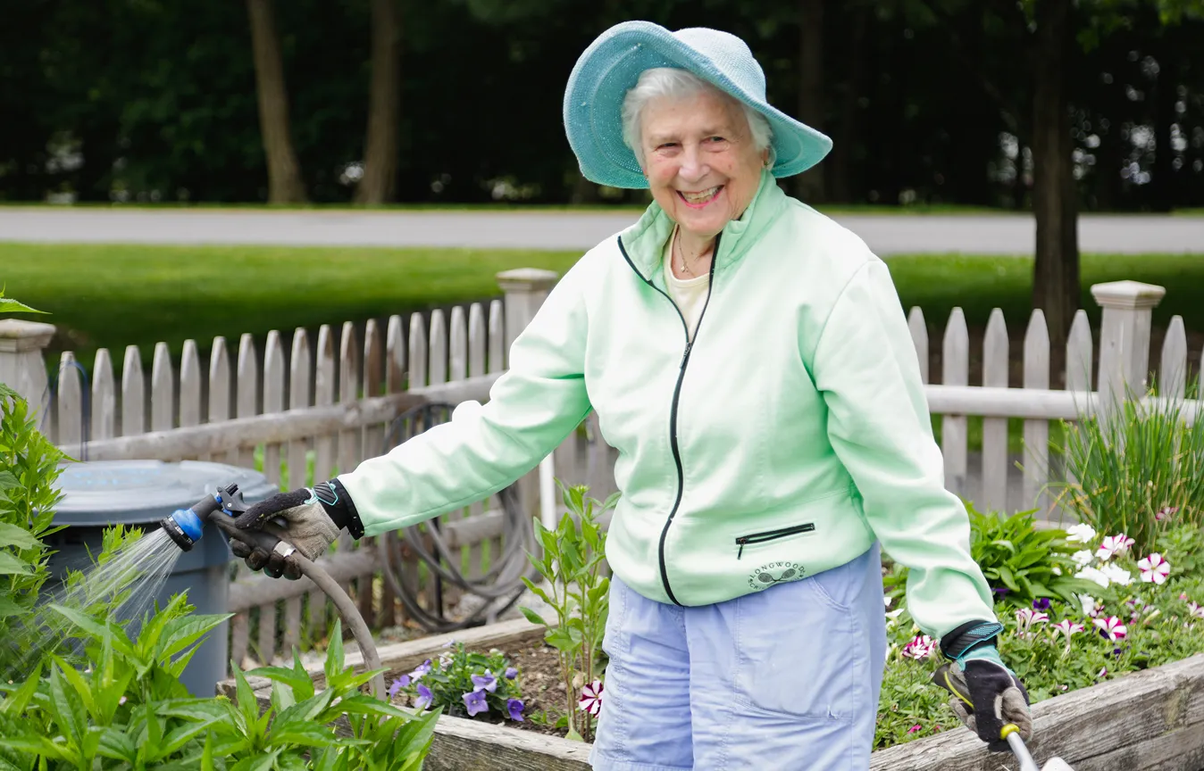 An older woman wearing a light green jacket and sunhat smiles while watering flowers in a raised garden bed. Surrounded by greenery and a wooden fence, she enjoys a peaceful moment in the community garden.