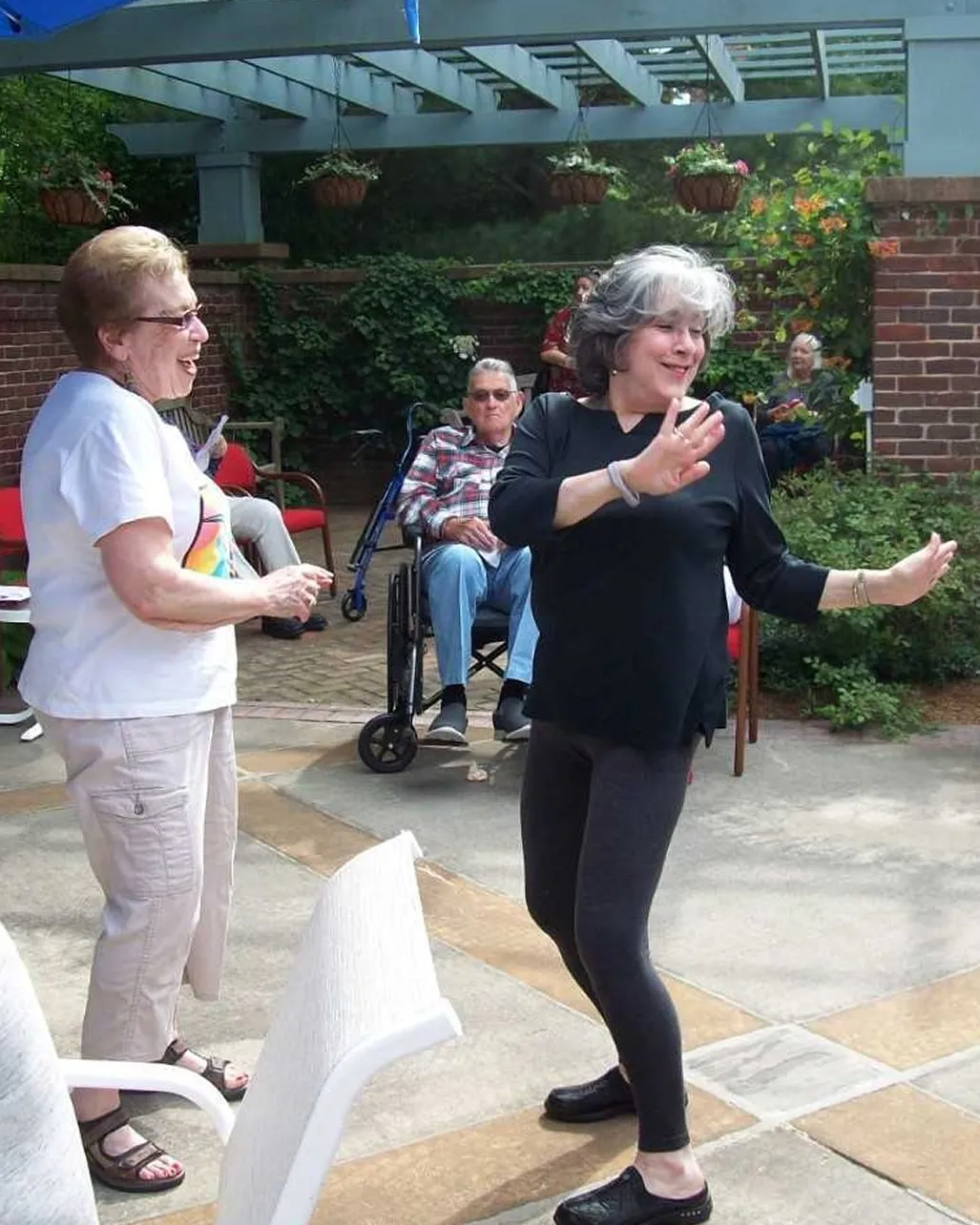 Two women smile and dance together on a sunny patio surrounded by greenery and hanging plants. Other residents watch and relax nearby, creating a joyful, community-centered atmosphere.