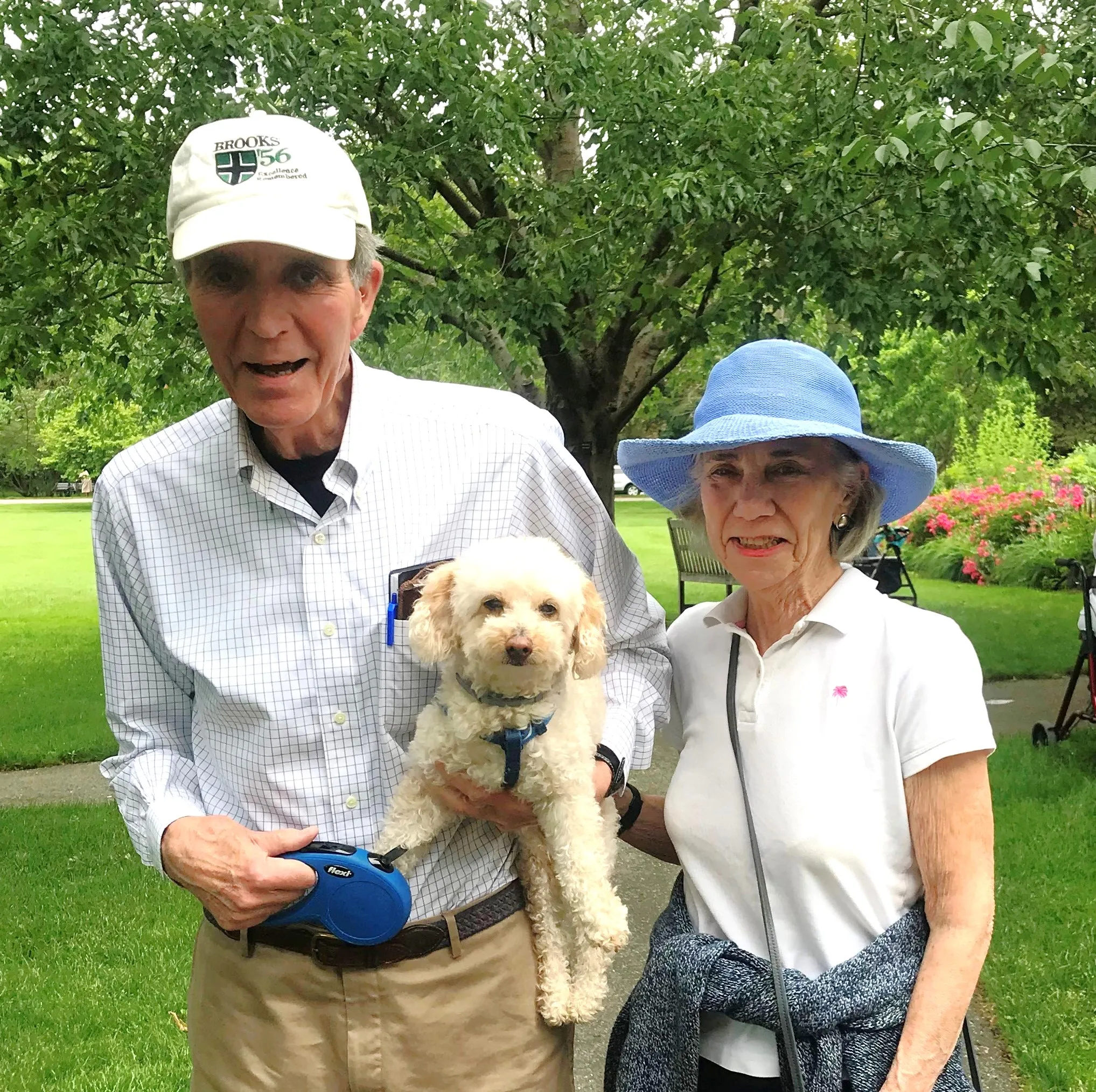 A smiling Laurelmead couple pauses on a lush green path with their small white dog in tow, enjoying a peaceful day outdoors. The moment captures the warmth of companionship and the joy of pet-friendly living.