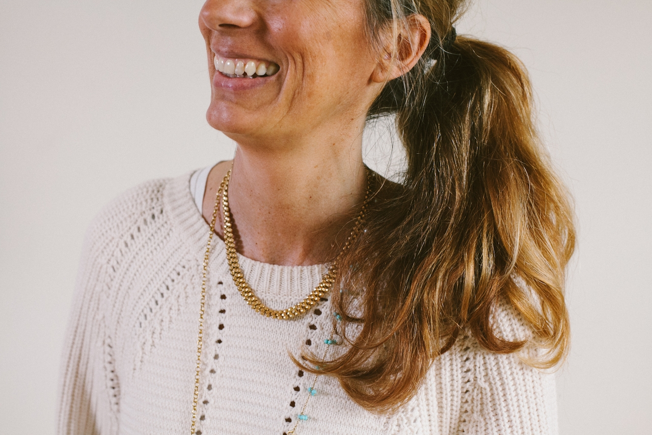 Smiling woman with a long ponytail wearing a white knit sweater and layered necklaces