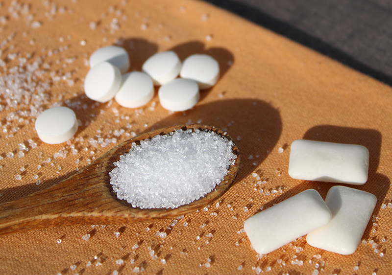 Xylitol crystals on a wooden spoon beside pieces of sugar-free gum