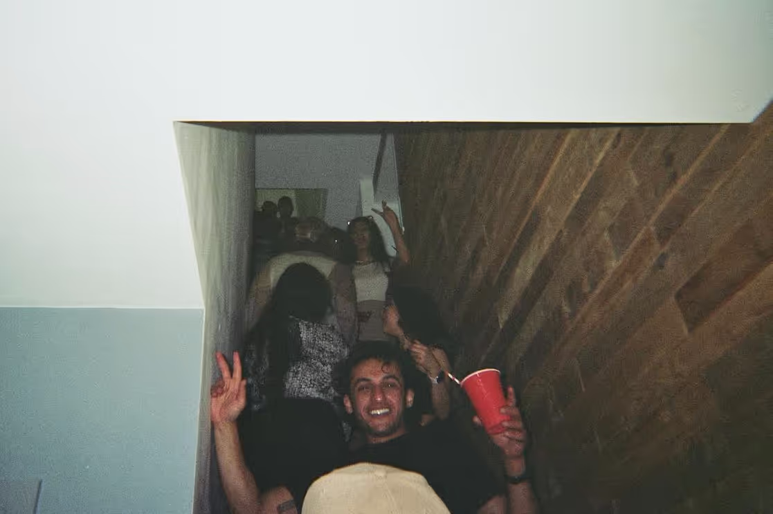 A young man smiling while the crowd goes upstairs at Soirée's "Back To The Roots" house party in Brighton, UK
