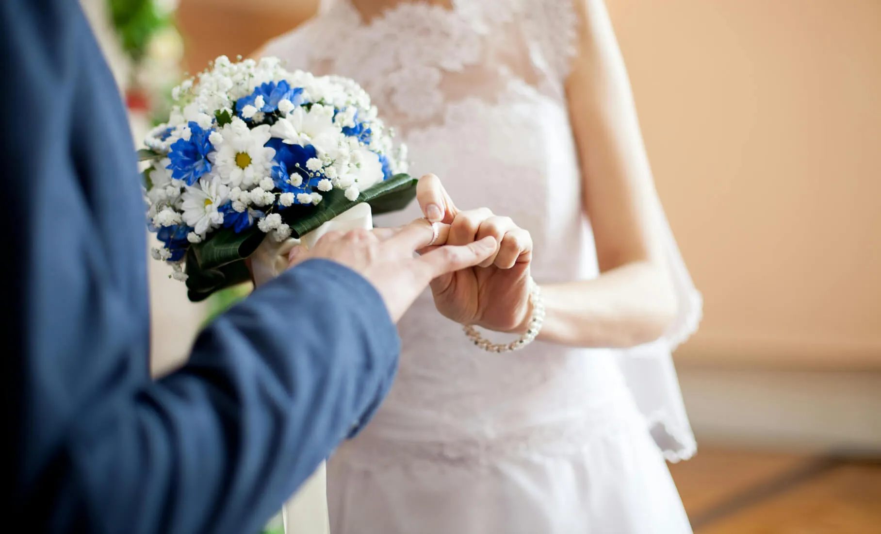 Bride placing a wedding ring on groom's finger, holding a bouquet of white and blue flowers.