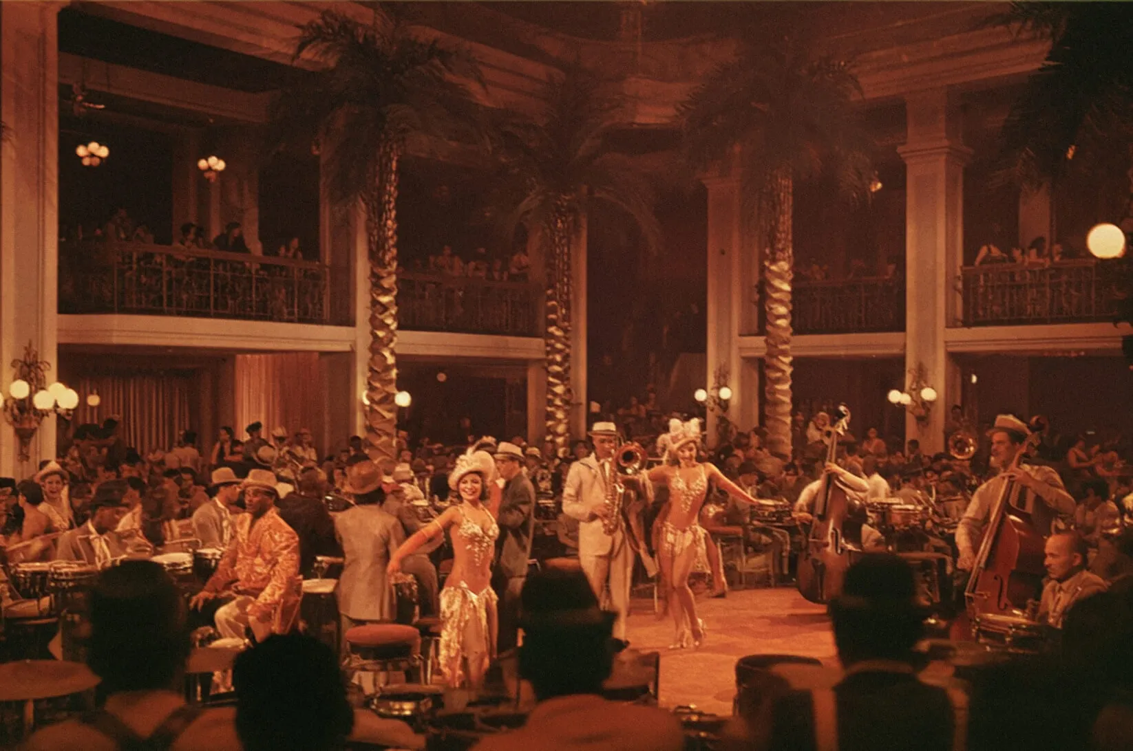 Vintage nightclub scene with two female dancers in sparkling costumes performing on a stage surrounded by musicians and a seated audience.