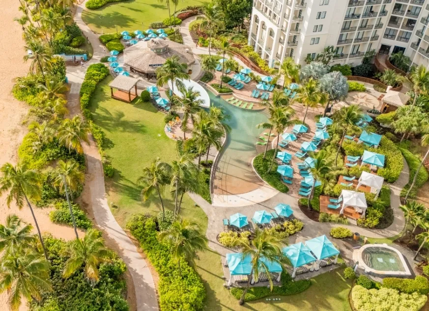 Aerial view of a resort pool area with blue umbrellas, palm trees, lounge chairs, huts, and pathways surrounded by greenery.