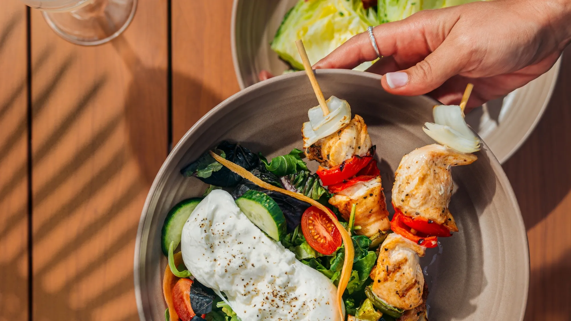Hand holding a bowl with grilled chicken skewers, fresh mixed greens, cucumbers, cherry tomatoes, shredded carrot, and creamy dressing.