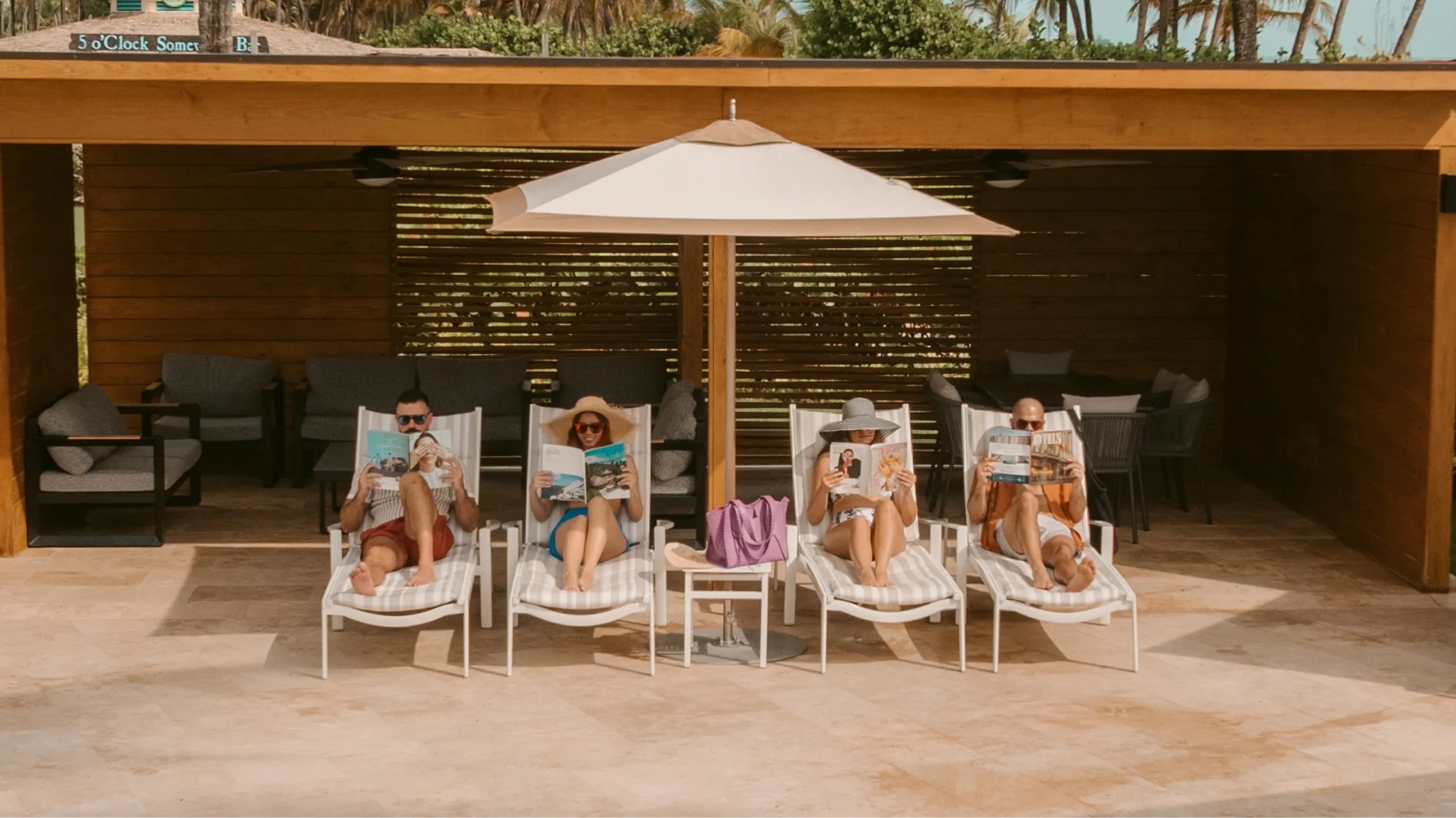 Four people lounging on striped beach chairs under a large umbrella, reading magazines by a wooden cabana with outdoor seating.
