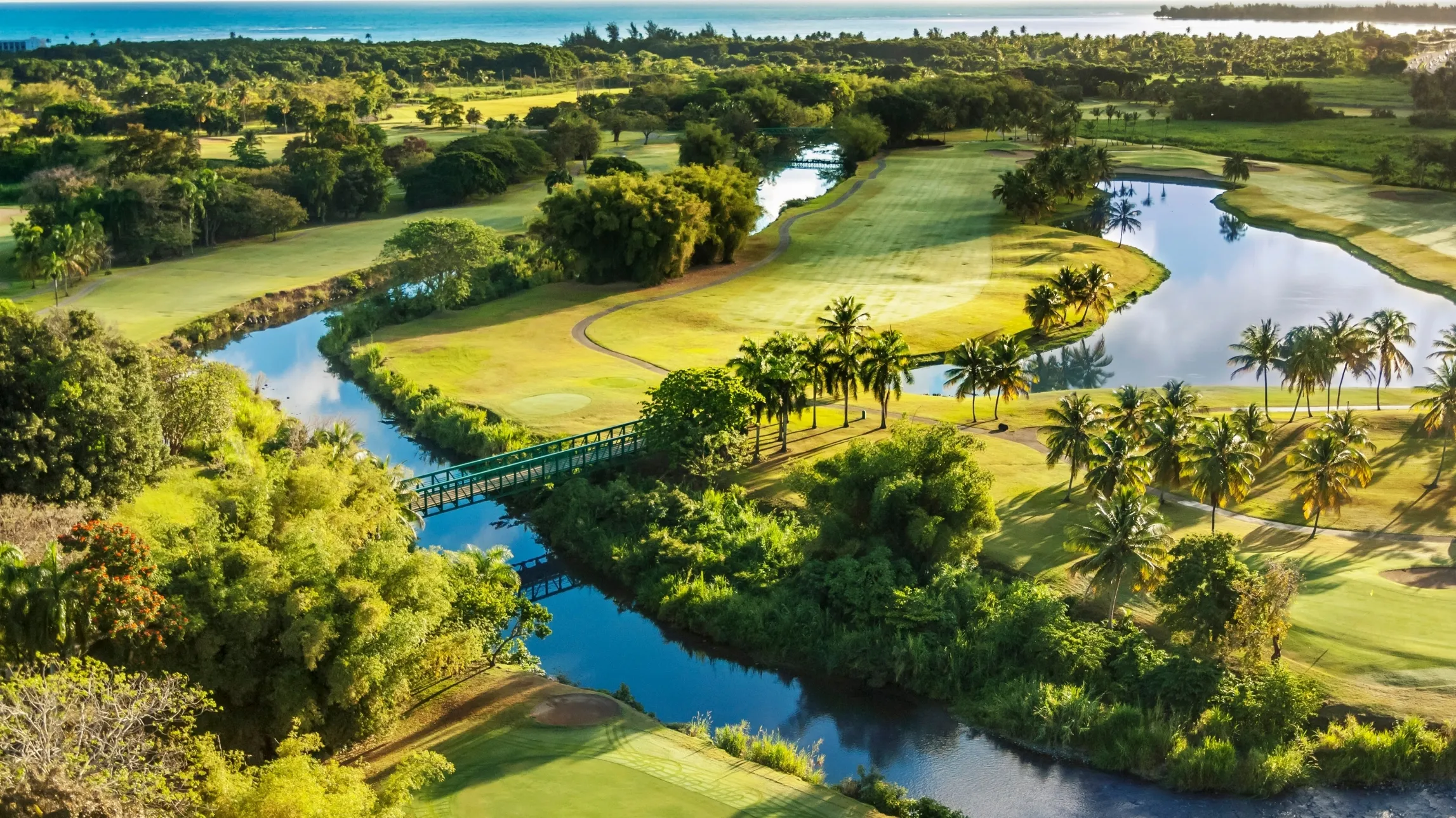 Aerial view of a golf course with palm trees, a small river, bridges, and the ocean in the background.