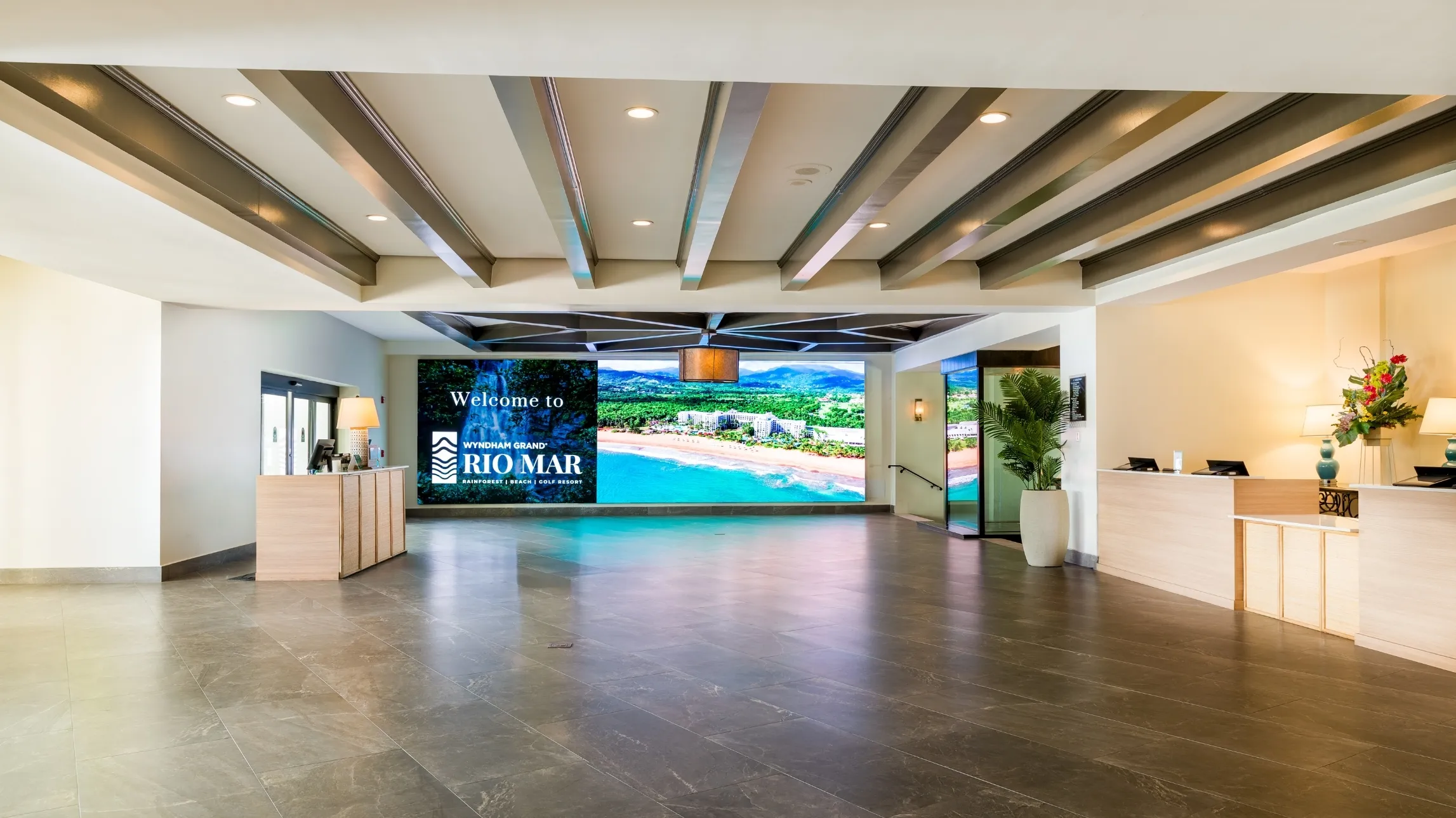Spacious hotel lobby with a large digital welcome sign for Wyndham Grand Rio Mar, two light wood reception desks, potted plant, and ceiling beams.