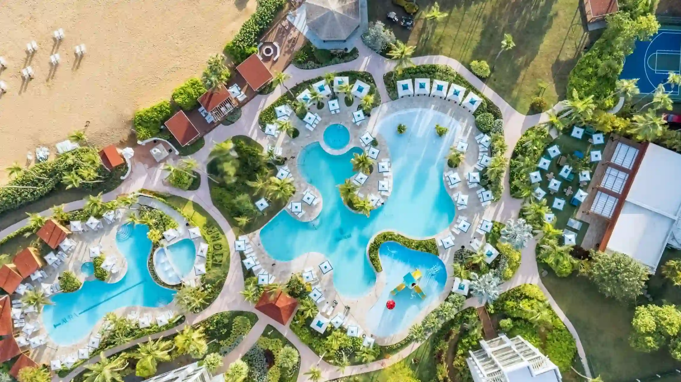 Aerial view of a resort pool area with multiple irregularly shaped pools surrounded by lounge chairs, umbrellas, cabanas, palm trees, and landscaped gardens.