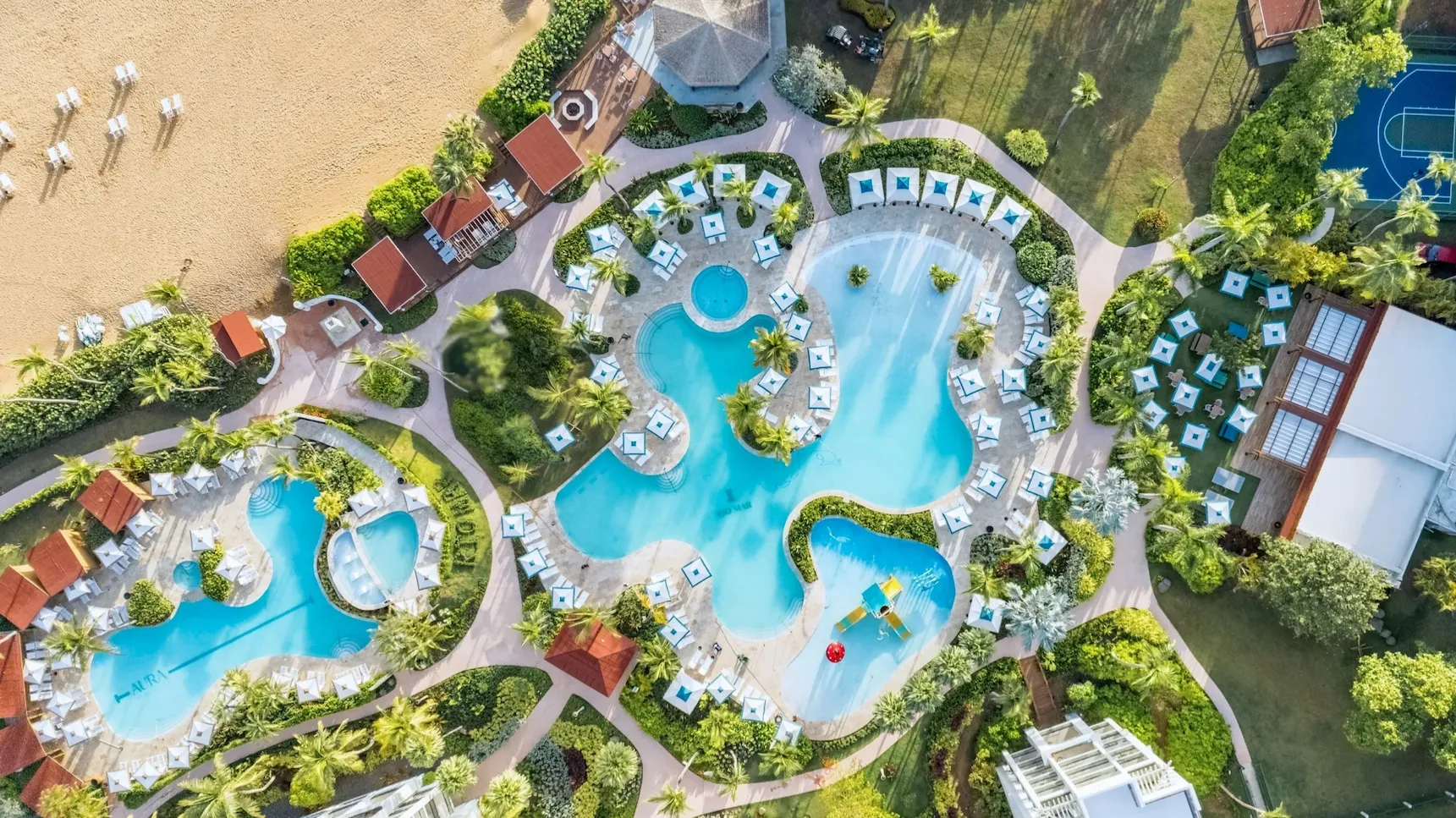 Aerial view of a resort pool area with multiple irregularly shaped pools surrounded by lounge chairs, umbrellas, cabanas, palm trees, and landscaped gardens.