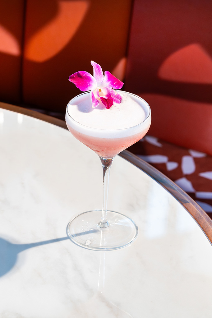 A pink cocktail in a martini glass topped with white foam and a pink plumeria garnish