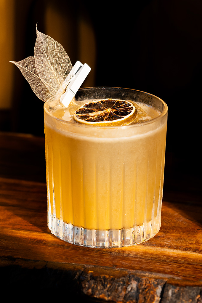 A pink cocktail in a martini glass topped with white foam and a pink plumeria garnish