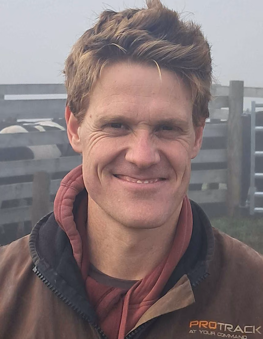 Man with short, tousled brown hair smiling, wearing a brown jacket and maroon hoodie, standing outdoors near wooden fences in a foggy setting.