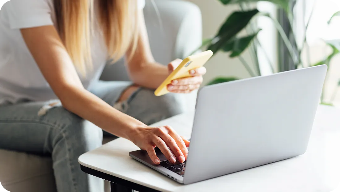 Person using a laptop while holding a smartphone at a desk.