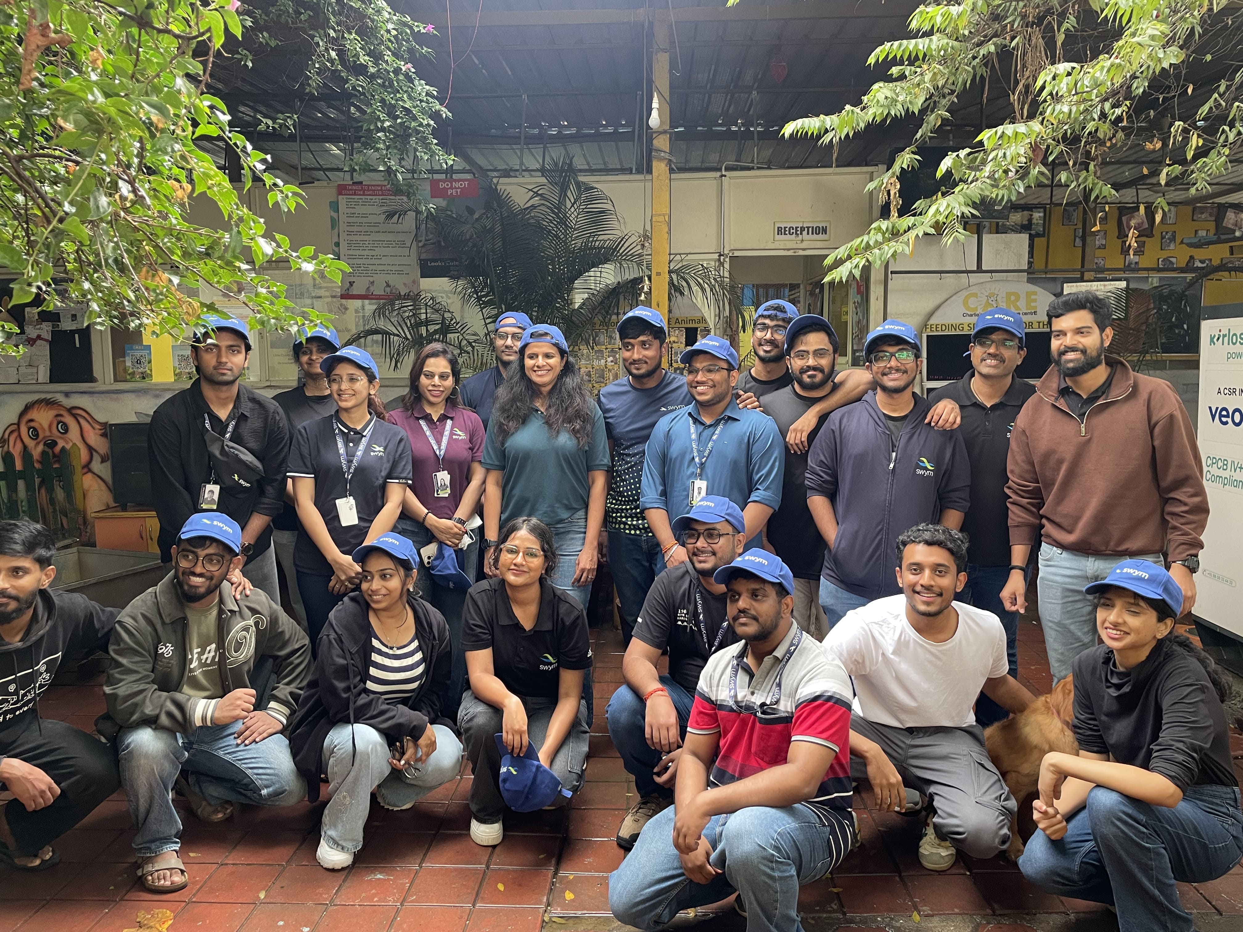 Group of 20 people, mostly young adults, posing in an indoor garden area with plants and signage including a dog illustration and reception sign, many wearing blue caps and some with company lanyards.